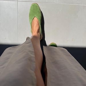 May include: A pair of bright green suede slip-on shoes. The shoes have a pointed toe and a low heel. The image is taken from a low angle, showing the shoes and part of the person's legs and a beige skirt.