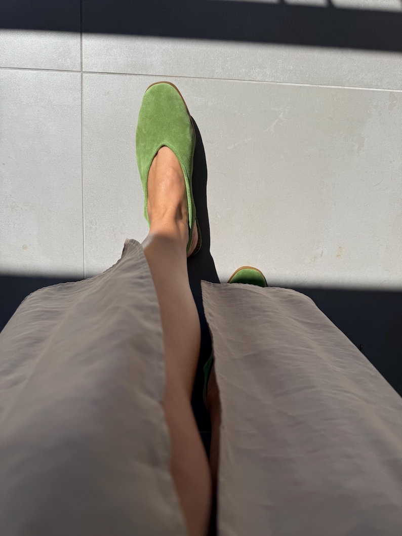 May include: A pair of bright green suede slip-on shoes. The shoes have a pointed toe and a low heel. The image is taken from a low angle, showing the shoes and part of the person's legs and a beige skirt.