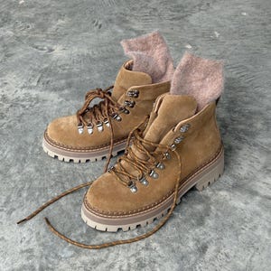 Op de afbeelding: Een paar bruin suède wandelschoenen met bruine veters en metalen vetergaten. De laarzen hebben een dikke, gebroken witte zool en zijn gevuld met lichtbruine sokken. Ontworpen voor buitenactiviteiten.