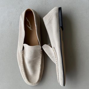 Puede incluir: Un par de mocasines de ante beige claro. Los zapatos tienen una puntera redondeada, detalles de costura y un tac&oacute;n bajo. El interior de un zapato muestra el nombre de la marca en letra cursiva. Estos mocasines est&aacute;n dise&ntilde;ados para la comodidad y el estilo.