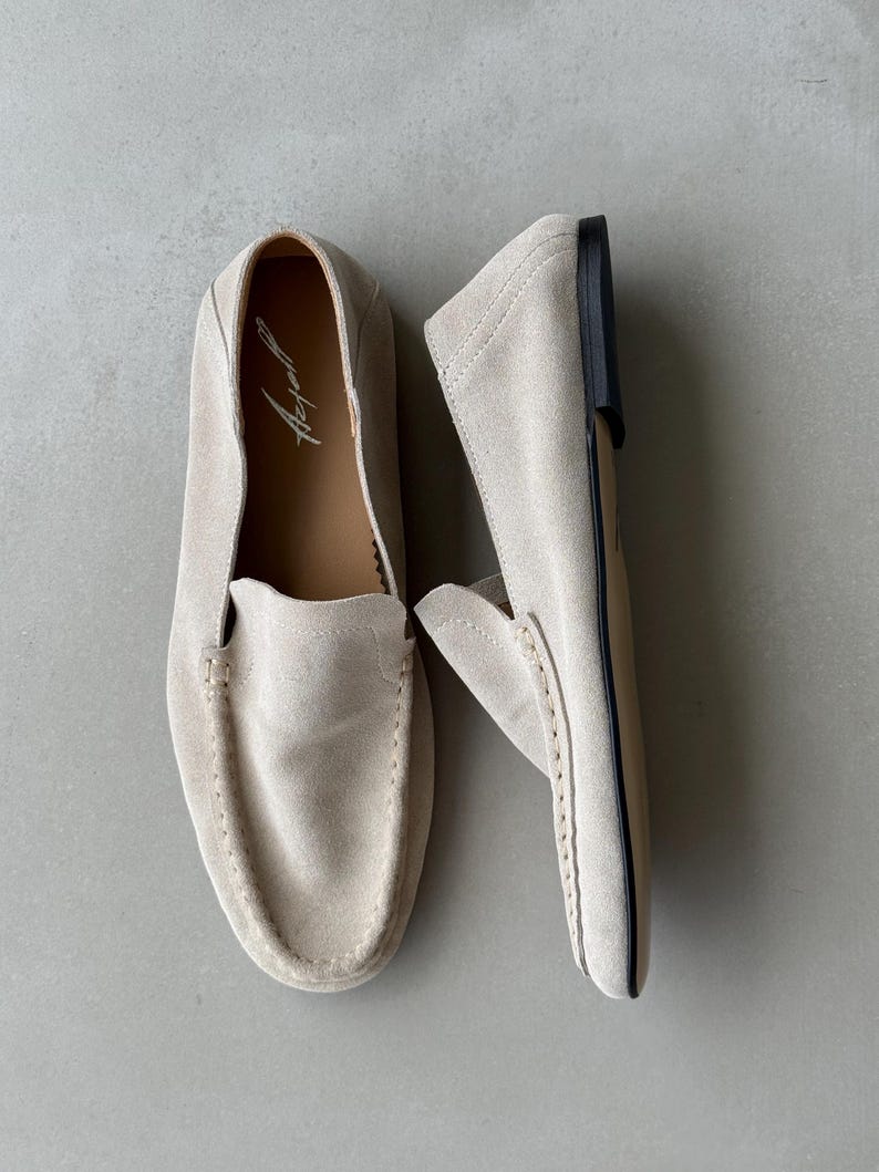 Puede incluir: Un par de mocasines de ante beige claro. Los zapatos tienen una puntera redondeada, detalles de costura y un tac&oacute;n bajo. El interior de un zapato muestra el nombre de la marca en letra cursiva. Estos mocasines est&aacute;n dise&ntilde;ados para la comodidad y el estilo.
