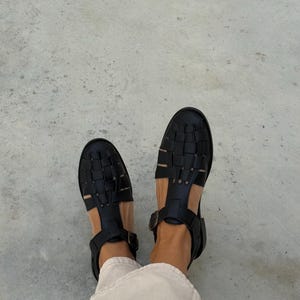 K&ouml;nnte beinhalten: Schwarze Ledersandalen mit gewebtem Design. Die Sandalen haben eine geschlossene Spitze und Kn&ouml;chelriemen mit Schnallen. Draufsicht.