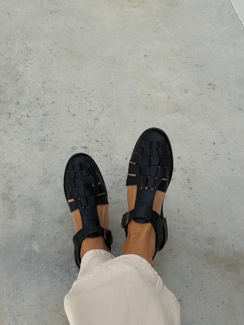 K&ouml;nnte beinhalten: Schwarze Ledersandalen mit gewebtem Design. Die Sandalen haben eine geschlossene Spitze und Kn&ouml;chelriemen mit Schnallen. Draufsicht.