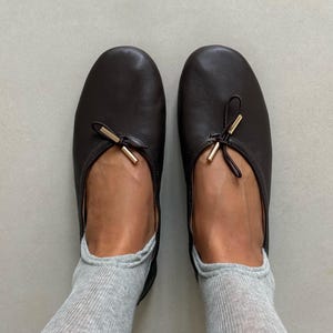 May include: A pair of dark brown leather ballet flats featuring a rounded toe and a decorative bow detail on the vamp. The bow is accented with small, cylindrical gold-tone metal tips. The shoes are worn with light gray knit socks.