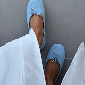 May include: A pair of light blue suede ballet flats featuring a rounded toe, a delicate drawstring tie at the vamp, and small gold-toned metal aglets at the ends of the ties. The shoes have a soft, matte texture and a simple, slip-on design.