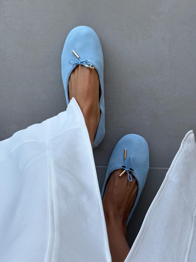 May include: A pair of light blue suede ballet flats featuring a rounded toe, a delicate drawstring tie at the vamp, and small gold-toned metal aglets at the ends of the ties. The shoes have a soft, matte texture and a simple, slip-on design.