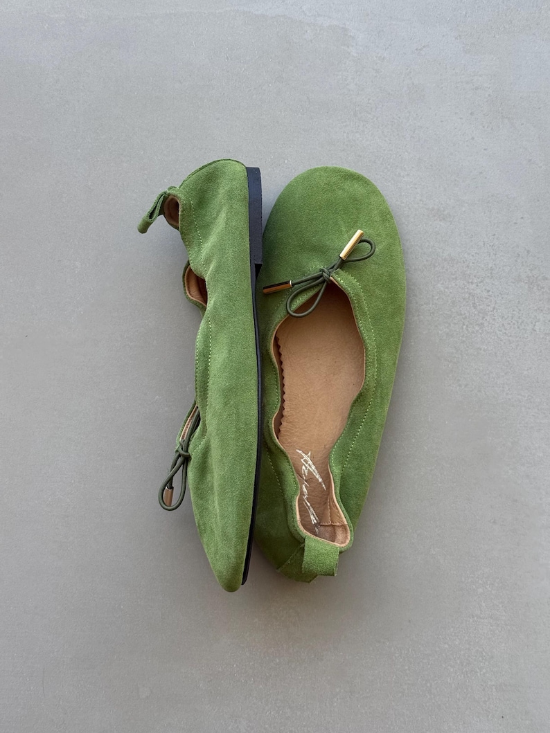 May include: A pair of bright green suede ballet flats featuring a rounded toe, a thin drawstring tie with gold-toned metal aglets at the front, a tan interior lining, and a small pull tab at the heel. The shoes have a flat black sole.