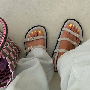 May include: Sandals with beige straps and gold buckles. The sandals have a black sole and are worn with light-colored pants. The toenails are painted yellow. A pink, green, and brown woven basket is visible on the left.