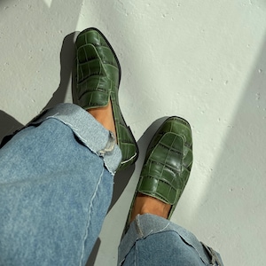 Könnte beinhalten: Ein Paar olivgrüne Slipper mit einer strukturierten Krokodilprägung und einer quadratischen Zehenpartie. Die Schuhe verfügen über ein gestepptes Nahtmuster auf dem Vorderblatt und werden mit einer hellblauen Jeans mit Umschlag getragen.
