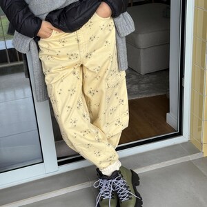 May include: A person wearing light yellow cargo pants with a floral pattern, a black puffer jacket, and olive green hiking boots with black soles and white and black laces. A gray sweater is draped over the shoulders.