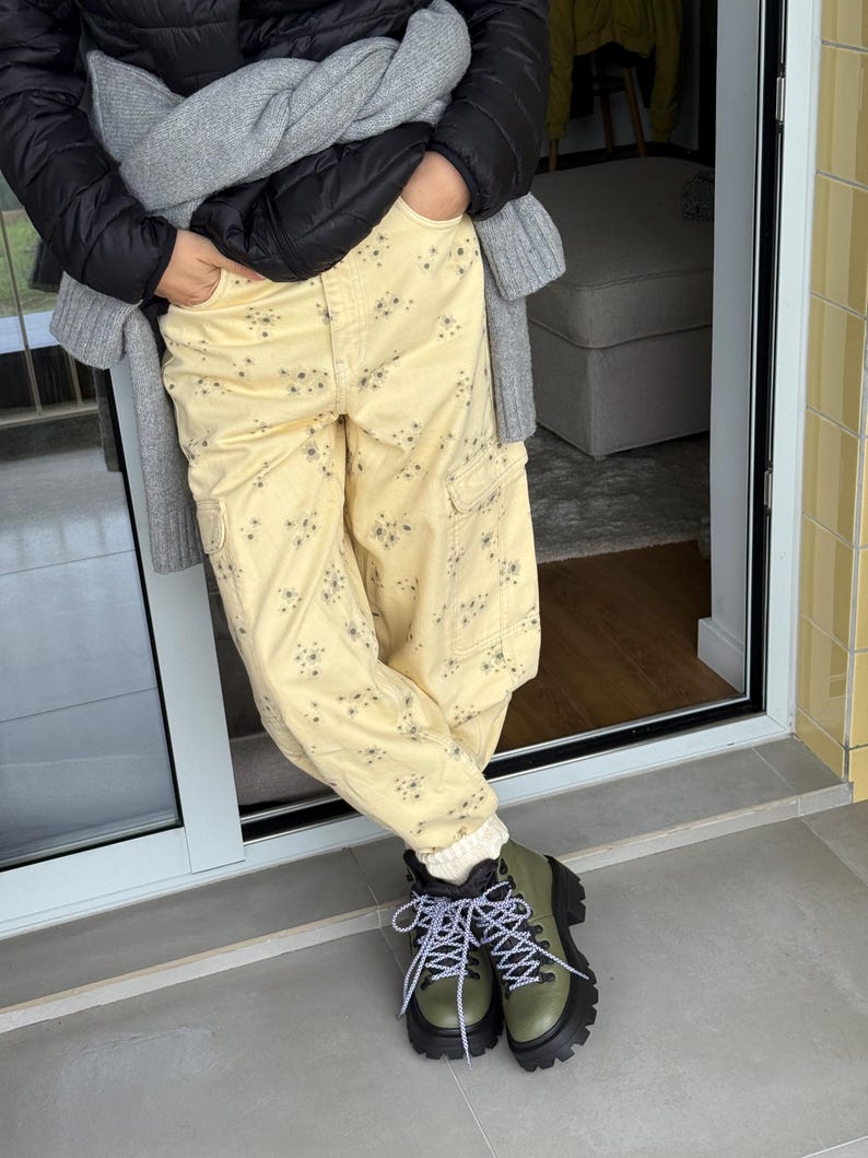 May include: A person wearing light yellow cargo pants with a floral pattern, a black puffer jacket, and olive green hiking boots with black soles and white and black laces. A gray sweater is draped over the shoulders.