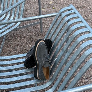 May include: A pair of gray suede boat shoes with thick black soles and light brown stitching. The shoes are laced up and have a classic boat shoe design. They are resting on a metal bench.