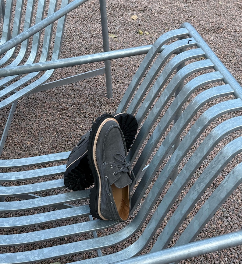May include: A pair of gray suede boat shoes with thick black soles and light brown stitching. The shoes are laced up and have a classic boat shoe design. They are resting on a metal bench.