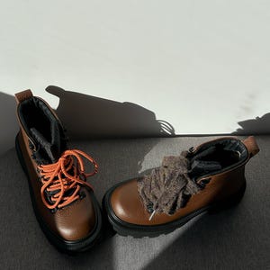 May include: A pair of brown leather hiking boots with black soles and orange laces. The boots have a rugged design, suitable for outdoor activities. The boots are displayed on a gray surface.