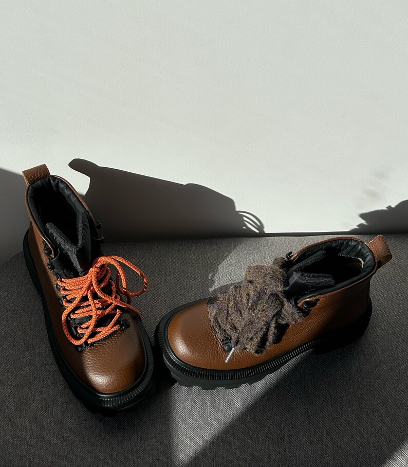 May include: A pair of brown leather hiking boots with black soles and orange laces. The boots have a rugged design, suitable for outdoor activities. The boots are displayed on a gray surface.