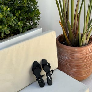 May include: A pair of black sandals with thin straps and small bows. The sandals have a minimalist design and are placed on a white surface. A potted plant and greenery are in the background.