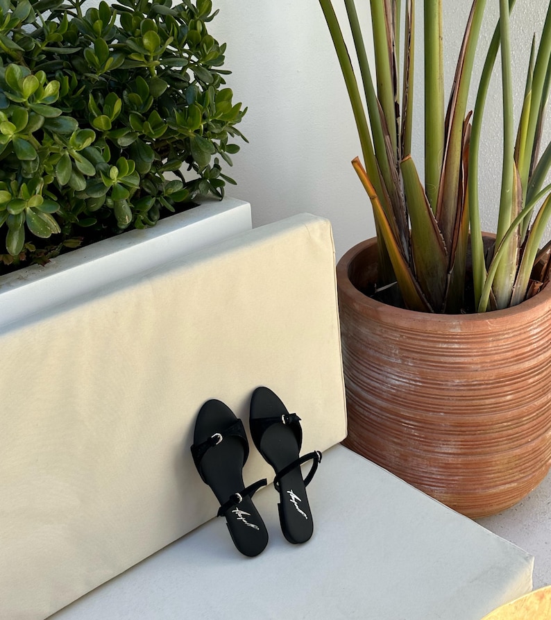 May include: A pair of black sandals with thin straps and small bows. The sandals have a minimalist design and are placed on a white surface. A potted plant and greenery are in the background.