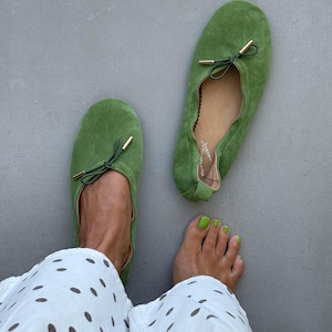 May include: A pair of grass green suede ballet flats featuring a rounded toe, a gathered topline, and a thin dark green drawstring bow detail with gold-toned metal aglets at the ends. The interior lining is tan and includes a signature logo.