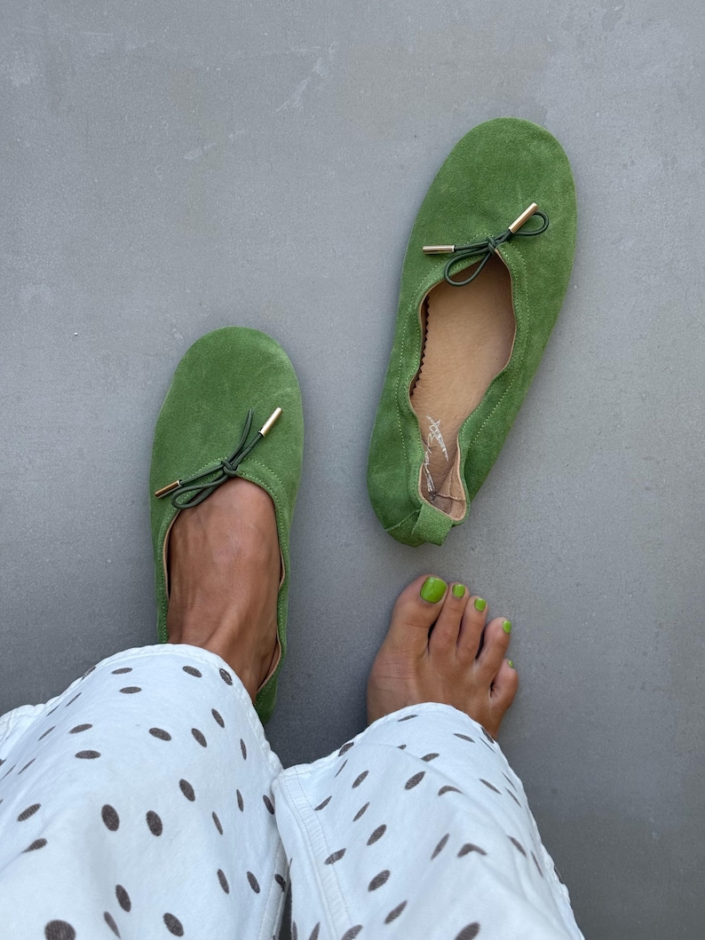 May include: A pair of grass green suede ballet flats featuring a rounded toe, a gathered topline, and a thin dark green drawstring bow detail with gold-toned metal aglets at the ends. The interior lining is tan and includes a signature logo.