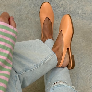 May include: A pair of peach-colored leather slingback flats with a rounded toe. The shoes have a low heel and a cutout detail on the vamp. The person is wearing light blue ripped jeans and a striped sleeve.