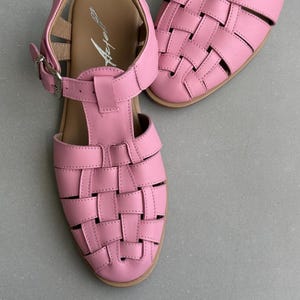 K&ouml;nnte beinhalten: Rosa Ledersandalen mit einem gewebten Design auf der Zehenpartie. Die Sandalen haben einen T-Riemen und einen Schnallenverschluss. Die Sohlen sind hellbraun, und die Sandalen stehen vor einem grauen Hintergrund.