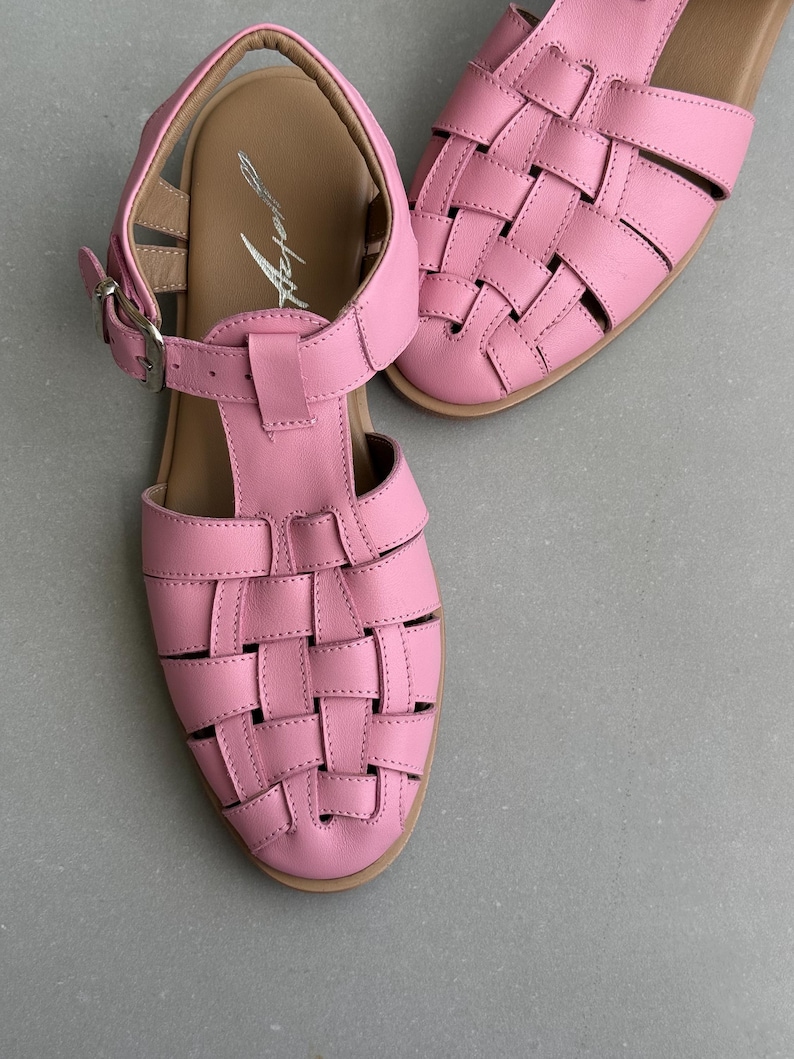 K&ouml;nnte beinhalten: Rosa Ledersandalen mit einem gewebten Design auf der Zehenpartie. Die Sandalen haben einen T-Riemen und einen Schnallenverschluss. Die Sohlen sind hellbraun, und die Sandalen stehen vor einem grauen Hintergrund.