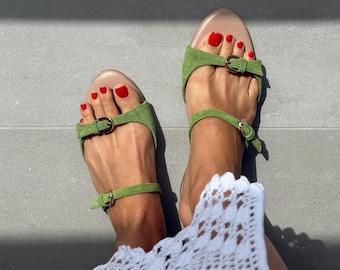 May include: A pair of green suede sandals with buckle closures. The sandals have a light brown footbed and are worn with a white lace dress. The person's toes are painted red.