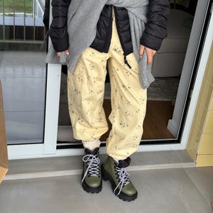 May include: A person wearing a black puffer jacket, gray sweater, and light yellow pants with a floral pattern. They are wearing olive green boots with black soles and white laces. The boots have a rugged, outdoor style.
