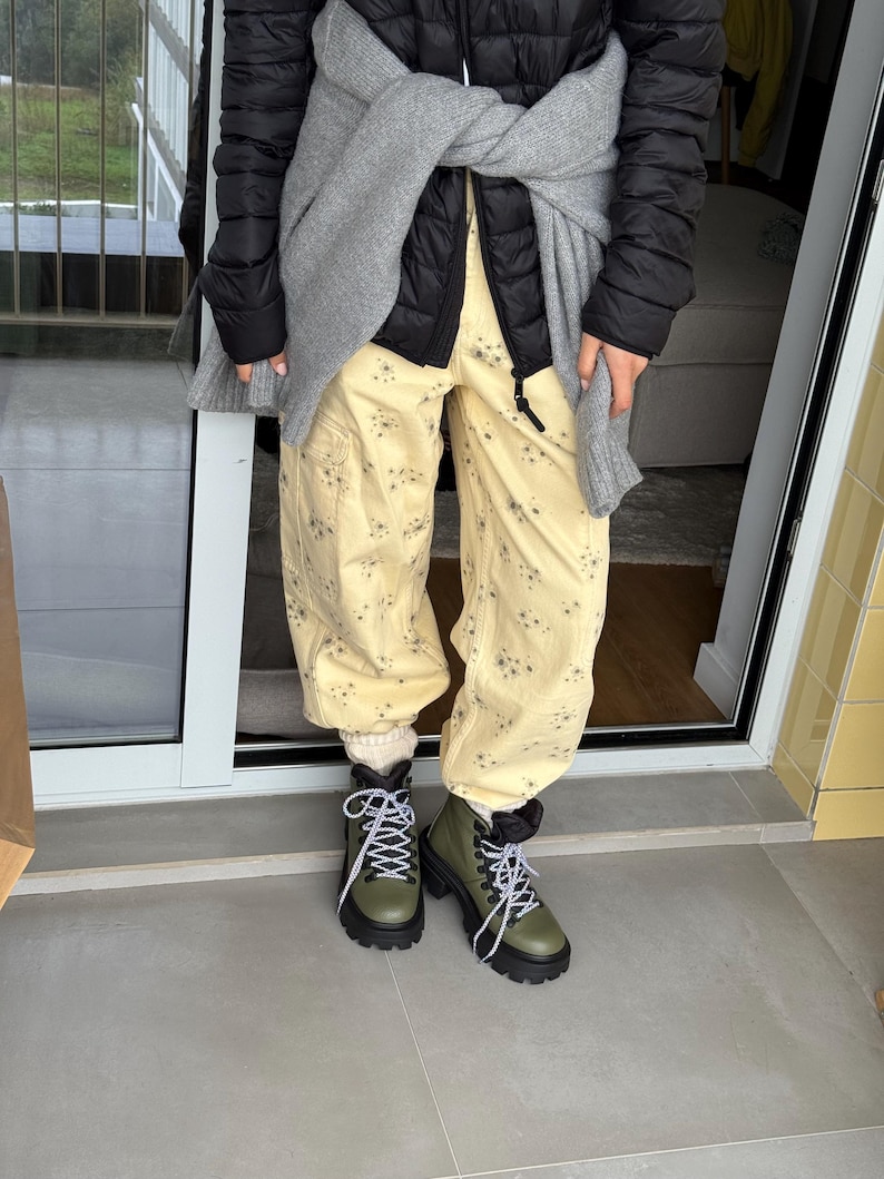 May include: A person wearing a black puffer jacket, gray sweater, and light yellow pants with a floral pattern. They are wearing olive green boots with black soles and white laces. The boots have a rugged, outdoor style.