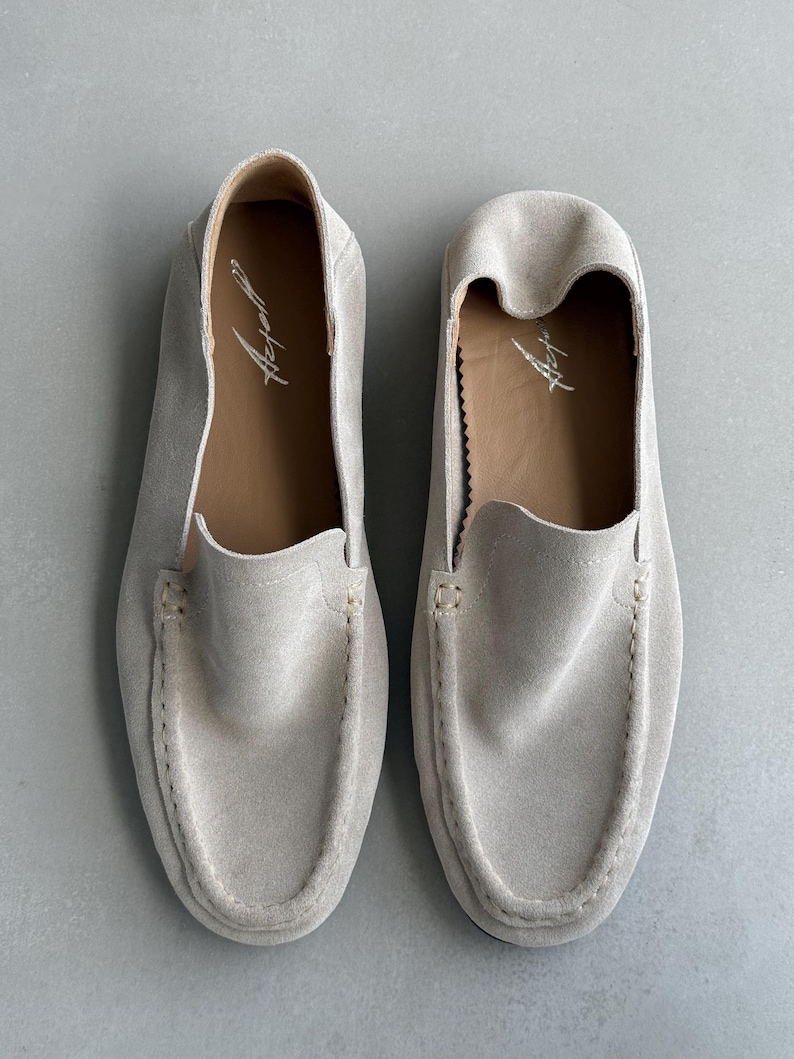 Puede incluir: Un par de mocasines de ante beige claro. Los zapatos tienen un dise&ntilde;o cl&aacute;sico sin cordones con punta redonda y empeine cosido. El interior es de color marr&oacute;n contrastante, con un logotipo escrito a mano. Los zapatos est&aacute;n planos sobre una superficie gris claro.