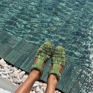 Puede incluir: Sandalias verdes con un dise&ntilde;o tejido. Las sandalias tienen un cierre de hebilla y est&aacute;n hechas de un material suave. Las sandalias se muestran cerca de una piscina con piedras blancas.