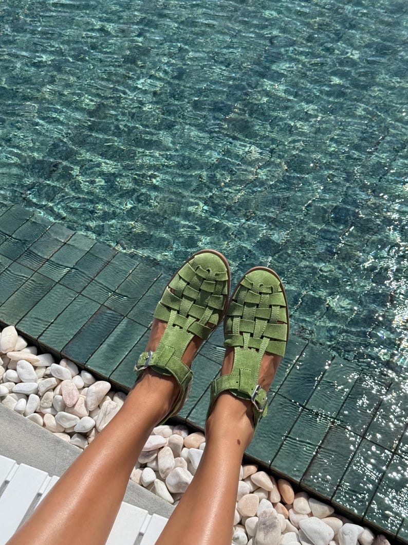 Puede incluir: Sandalias verdes con un dise&ntilde;o tejido. Las sandalias tienen un cierre de hebilla y est&aacute;n hechas de un material suave. Las sandalias se muestran cerca de una piscina con piedras blancas.