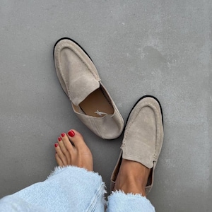 May include: Beige suede loafers with black soles. The shoes are positioned near a person's feet, with red nail polish on the toes, and light blue jeans. The loafers have a classic slip-on design, suitable for casual wear.