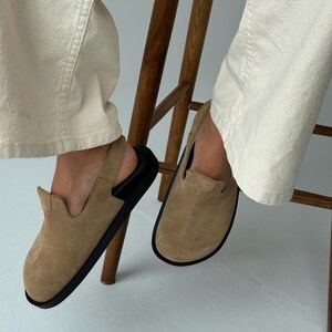 May include: Tan suede slip-on shoes with black soles. The shoes have a rounded toe and a low-profile design. The image shows the shoes being worn, with the wearer seated on a wooden chair.