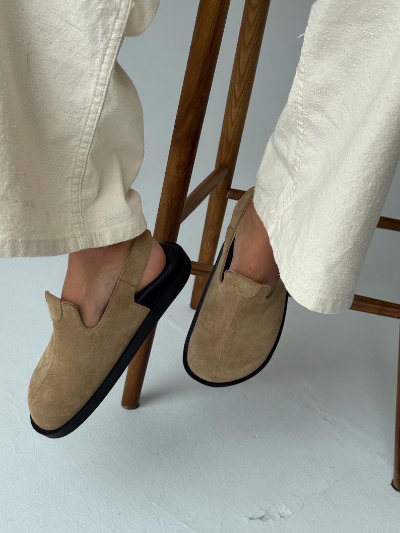 May include: Tan suede slip-on shoes with black soles. The shoes have a rounded toe and a low-profile design. The image shows the shoes being worn, with the wearer seated on a wooden chair.