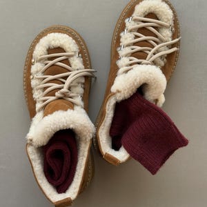 Könnte beinhalten: Ein Paar braune Wildleder-Wanderstiefel mit cremefarbenem Shearling-Futter und hellen Schnürsenkeln. Burgunderfarbene Socken sind in den Stiefeln zu sehen. Die Stiefel haben ein robustes Design, das für Outdoor-Aktivitäten geeignet ist.