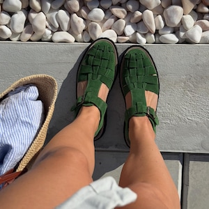 Puede incluir: Vista superior de sandalias verdes de cuero trenzado con hebillas. Las sandalias est&aacute;n sobre una superficie gris, con una bolsa de paja y tela a rayas en el fondo. Tambi&eacute;n se ven piedras blancas y un borde verde.
