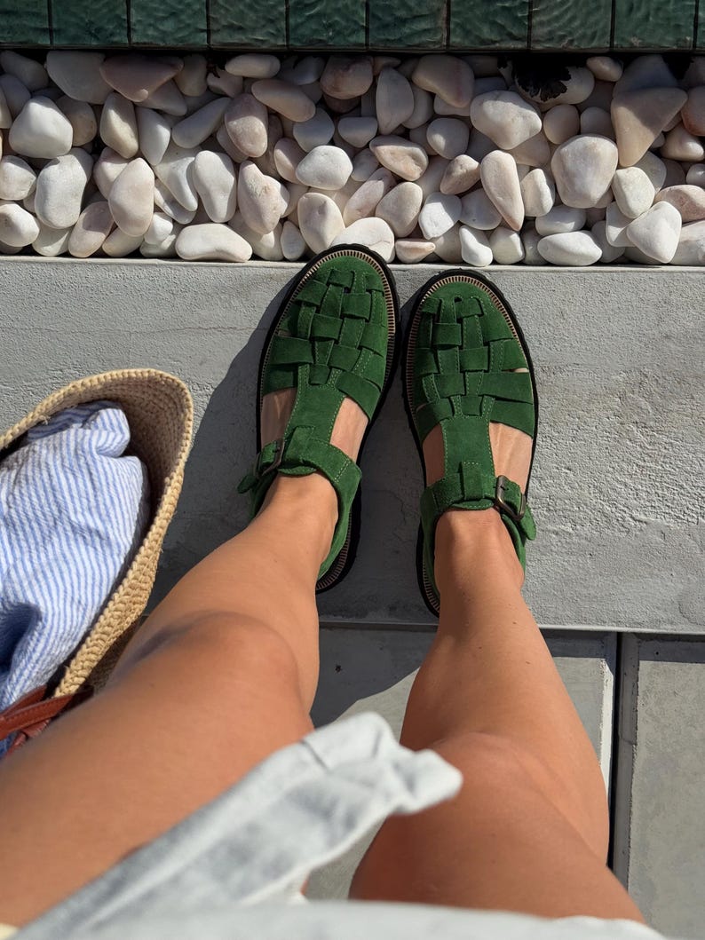 Puede incluir: Vista superior de sandalias verdes de cuero trenzado con hebillas. Las sandalias est&aacute;n sobre una superficie gris, con una bolsa de paja y tela a rayas en el fondo. Tambi&eacute;n se ven piedras blancas y un borde verde.