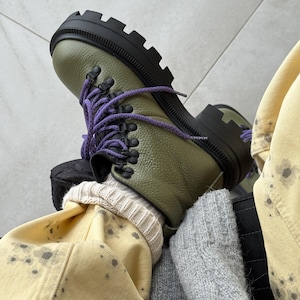 May include: Olive green leather hiking boots with black soles and purple laces. The boots have a chunky design and are laced up. The boots are worn with a cream-colored sock and yellow pants.