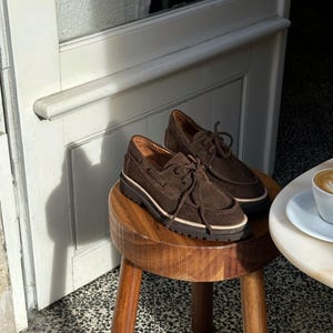 May include: A pair of brown suede boat shoes with laces, resting on a small wooden stool. The shoes have a thick, dark brown sole with a white stripe. The background includes a white door and a table with a coffee cup.