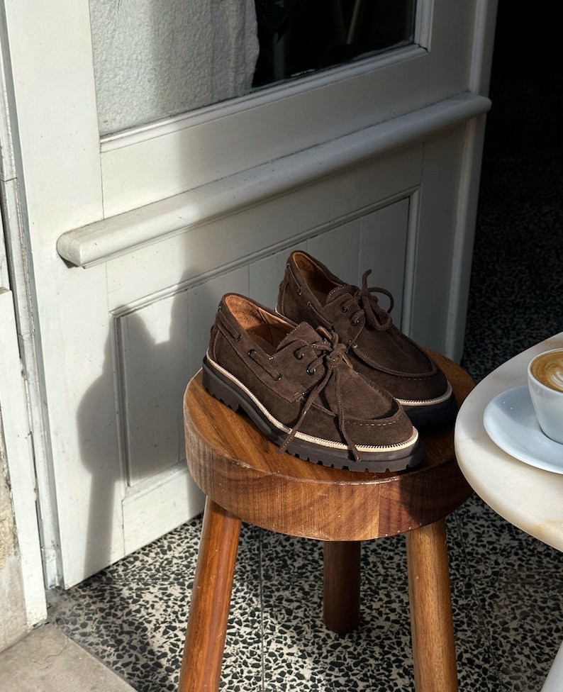 May include: A pair of brown suede boat shoes with laces, resting on a small wooden stool. The shoes have a thick, dark brown sole with a white stripe. The background includes a white door and a table with a coffee cup.