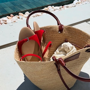 May include: A woven straw tote bag with dark brown leather handles and accents. Inside the bag are a pair of red sandals and a small drawstring pouch. The bag is sitting on a concrete surface.