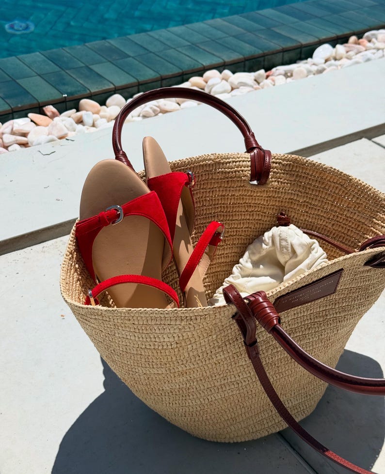 May include: A woven straw tote bag with dark brown leather handles and accents. Inside the bag are a pair of red sandals and a small drawstring pouch. The bag is sitting on a concrete surface.