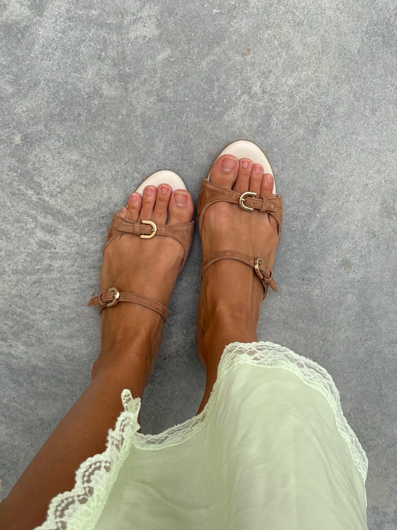 May include: Tan leather sandals with gold-colored buckles. The sandals have a toe strap and an ankle strap. The footbed is white. The sandals are worn with a light green dress.