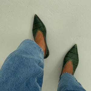 May include: A pair of dark green pointed-toe flats featuring a textured crocodile-embossed pattern and a deep V-shaped topline, worn with light blue denim jeans. The shoes have a sleek, low-profile silhouette with a structured finish.