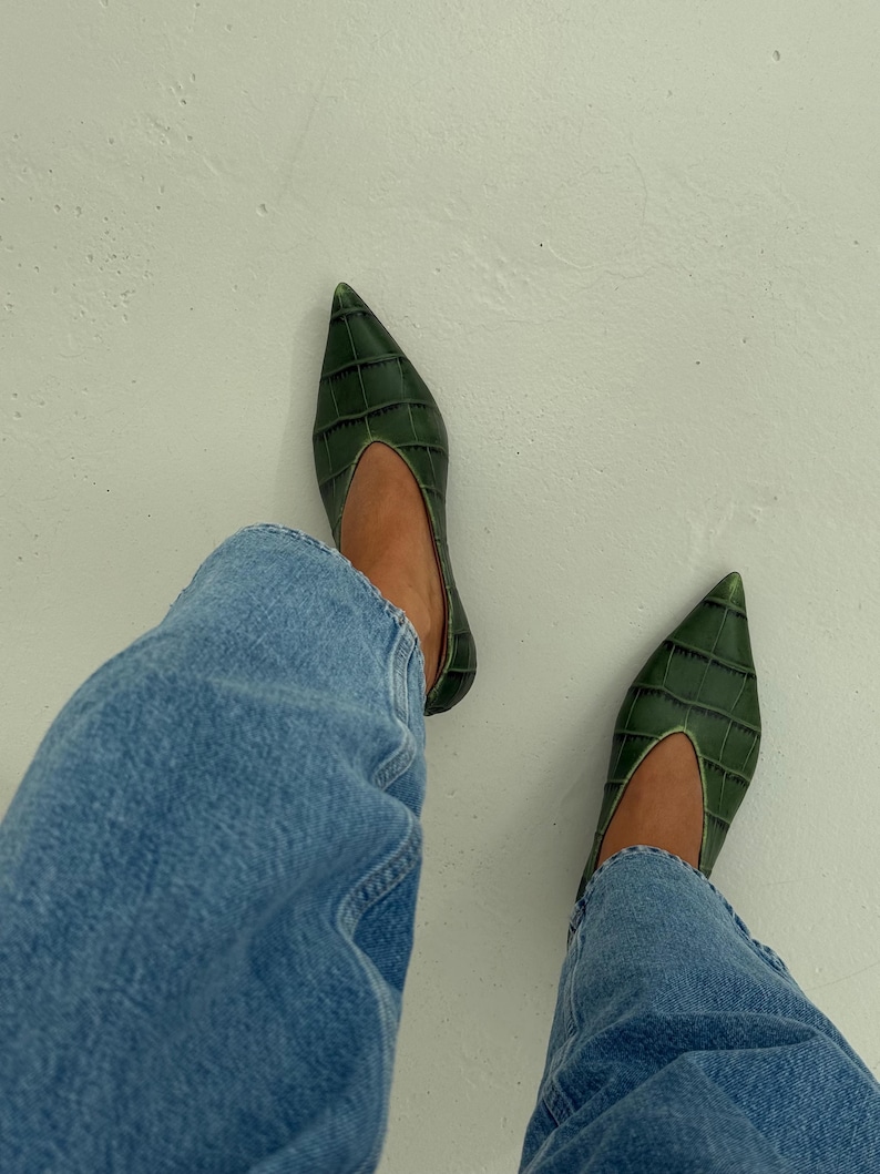 May include: A pair of dark green pointed-toe flats featuring a textured crocodile-embossed pattern and a deep V-shaped topline, worn with light blue denim jeans. The shoes have a sleek, low-profile silhouette with a structured finish.