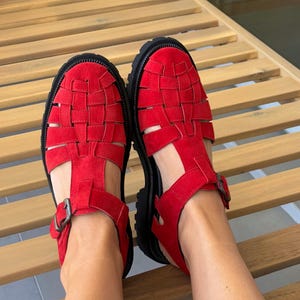 Puede incluir: Un par de sandalias rojas con un dise&ntilde;o tejido en la parte superior. Las sandalias tienen una suela negra y un cierre de hebilla. Los zapatos est&aacute;n hechos de un material similar al ante.
