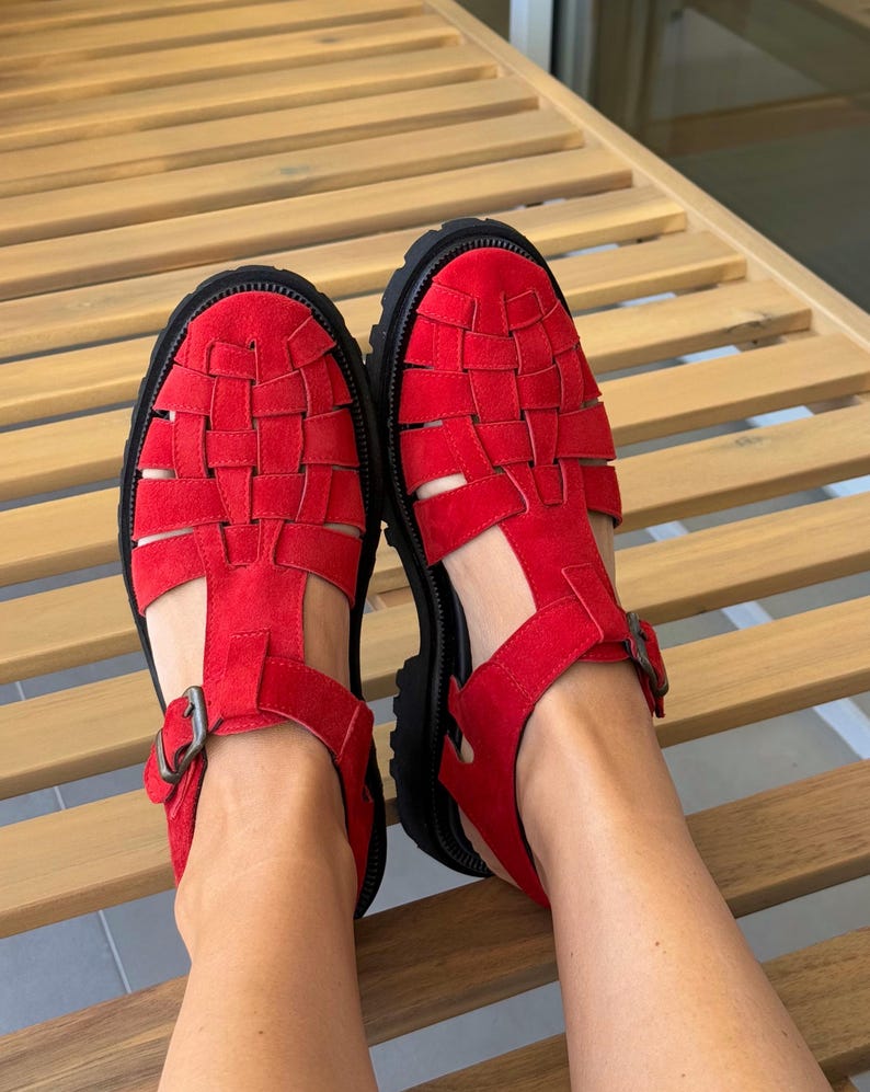 Puede incluir: Un par de sandalias rojas con un dise&ntilde;o tejido en la parte superior. Las sandalias tienen una suela negra y un cierre de hebilla. Los zapatos est&aacute;n hechos de un material similar al ante.