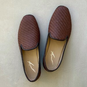May include: A pair of brown woven leather loafers. The shoes have a dark brown trim and a light-coloured interior with a signature. The loafers are displayed on a light grey surface.