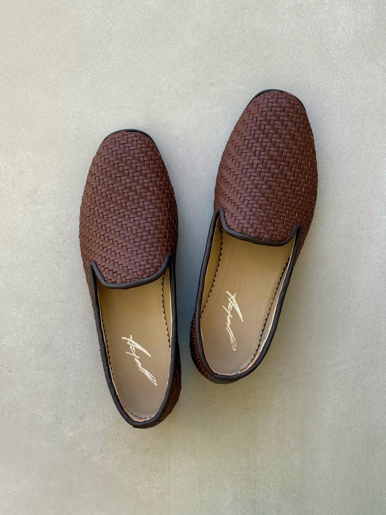 May include: A pair of brown woven leather loafers. The shoes have a dark brown trim and a light-coloured interior with a signature. The loafers are displayed on a light grey surface.